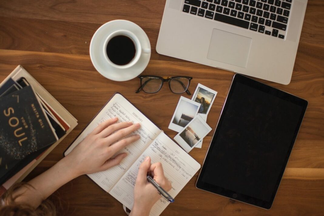 person writing in notebook with coffee, laptop, glasses, and pictures on table