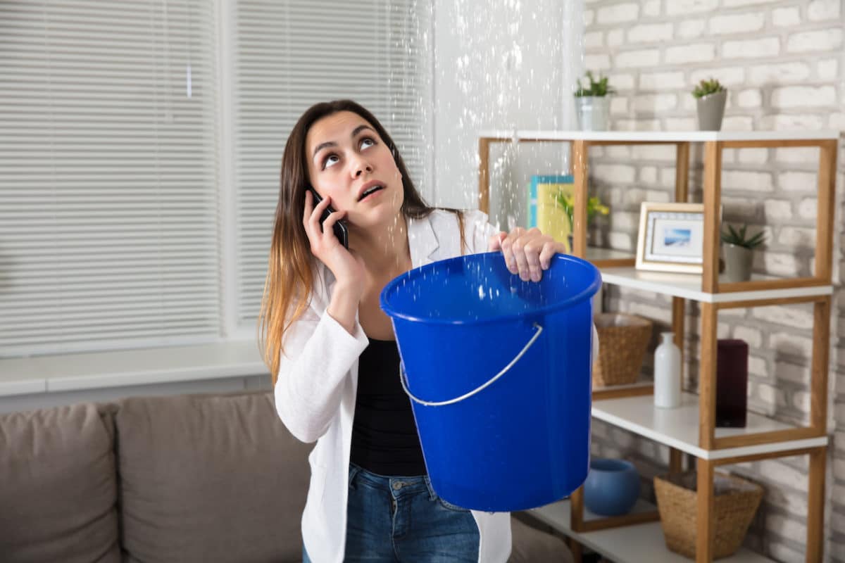 woman on the phone holding blue bucket to catch water leaking from ceiling 