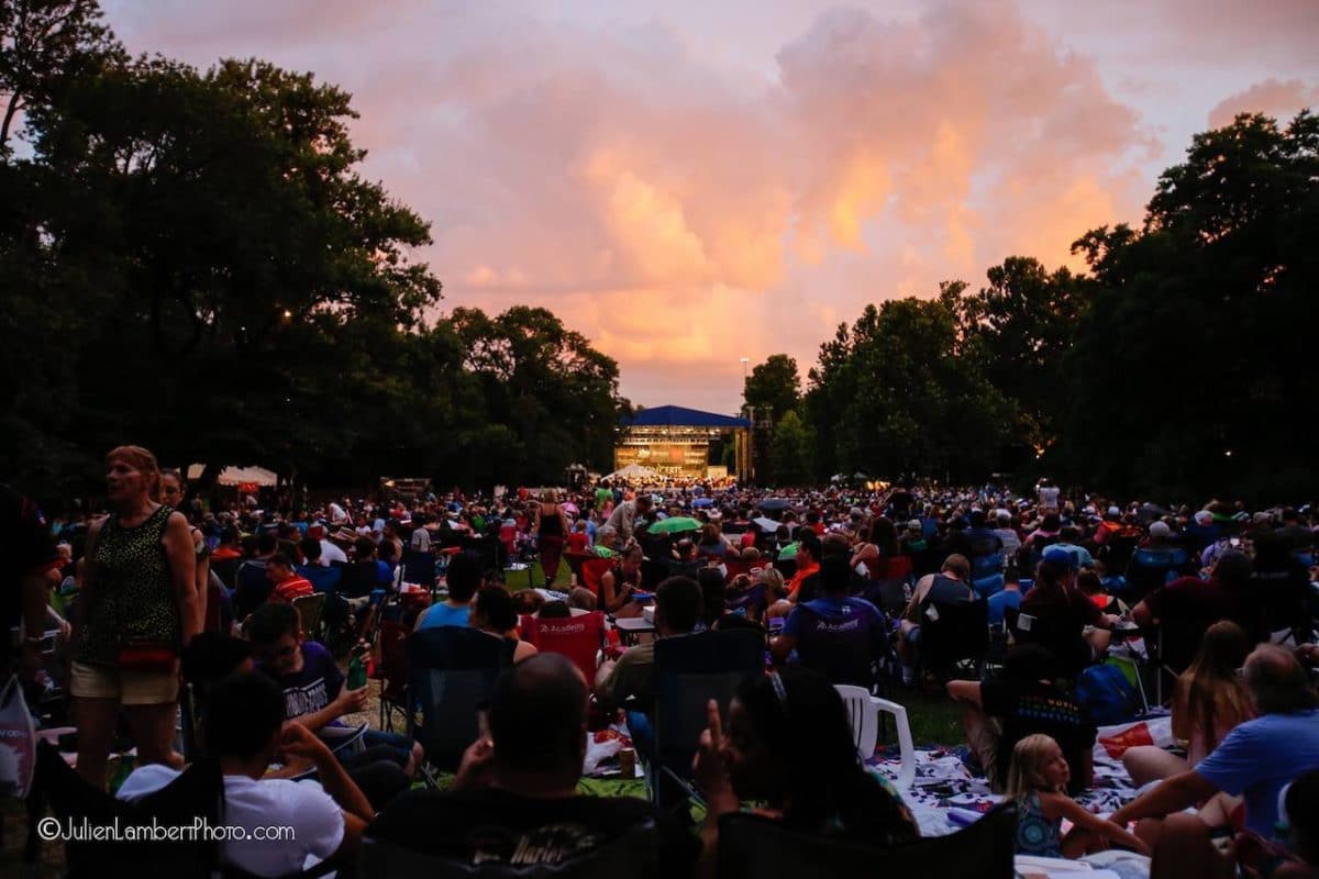 Crowd sitting on grass at sunset listening to music performer on stage