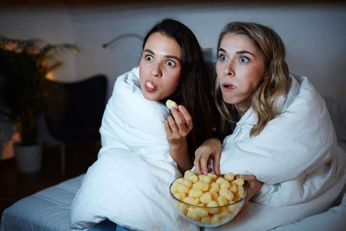 two women wrapped in blanket and eating snacks while watching a movie