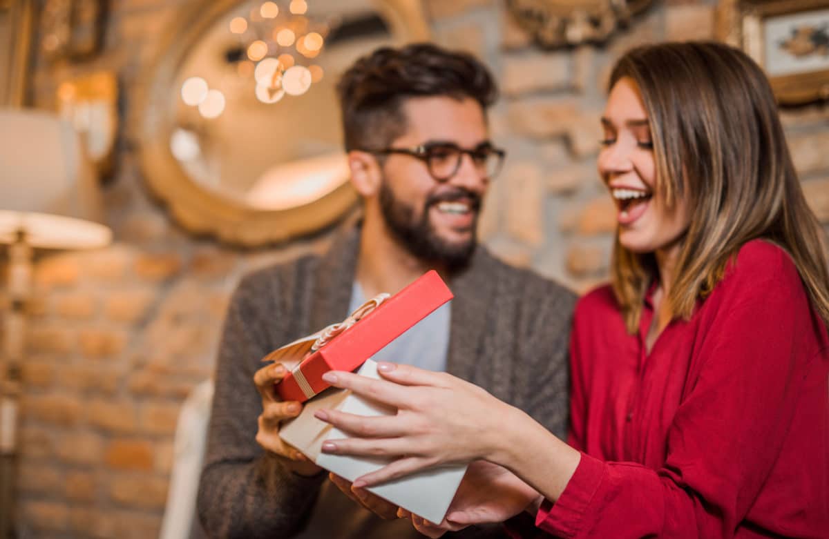 woman opening gift box and man sitting next to her watching