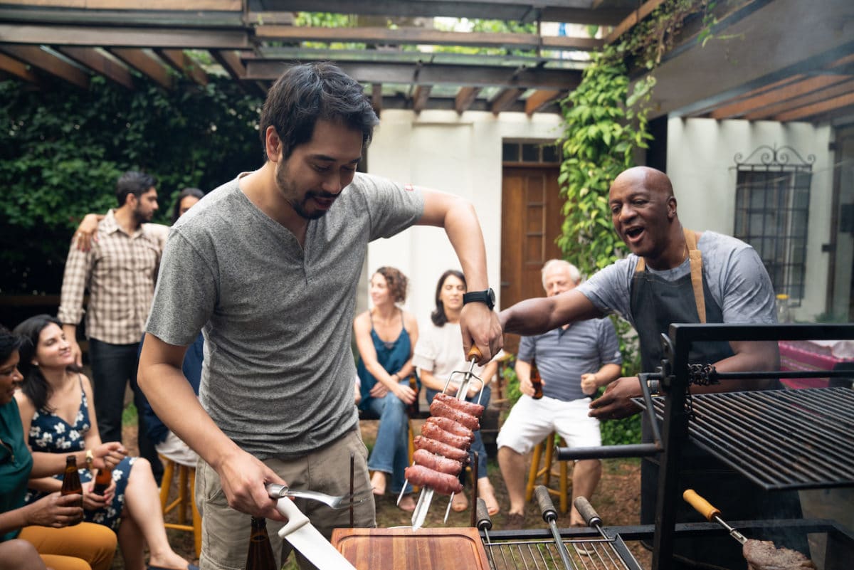 two men at bbq with other neighbors in background enjoying food and drinks