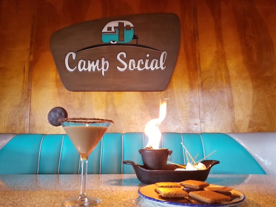 cocktail on table and s'mores items next to it with Camp Social sign in background