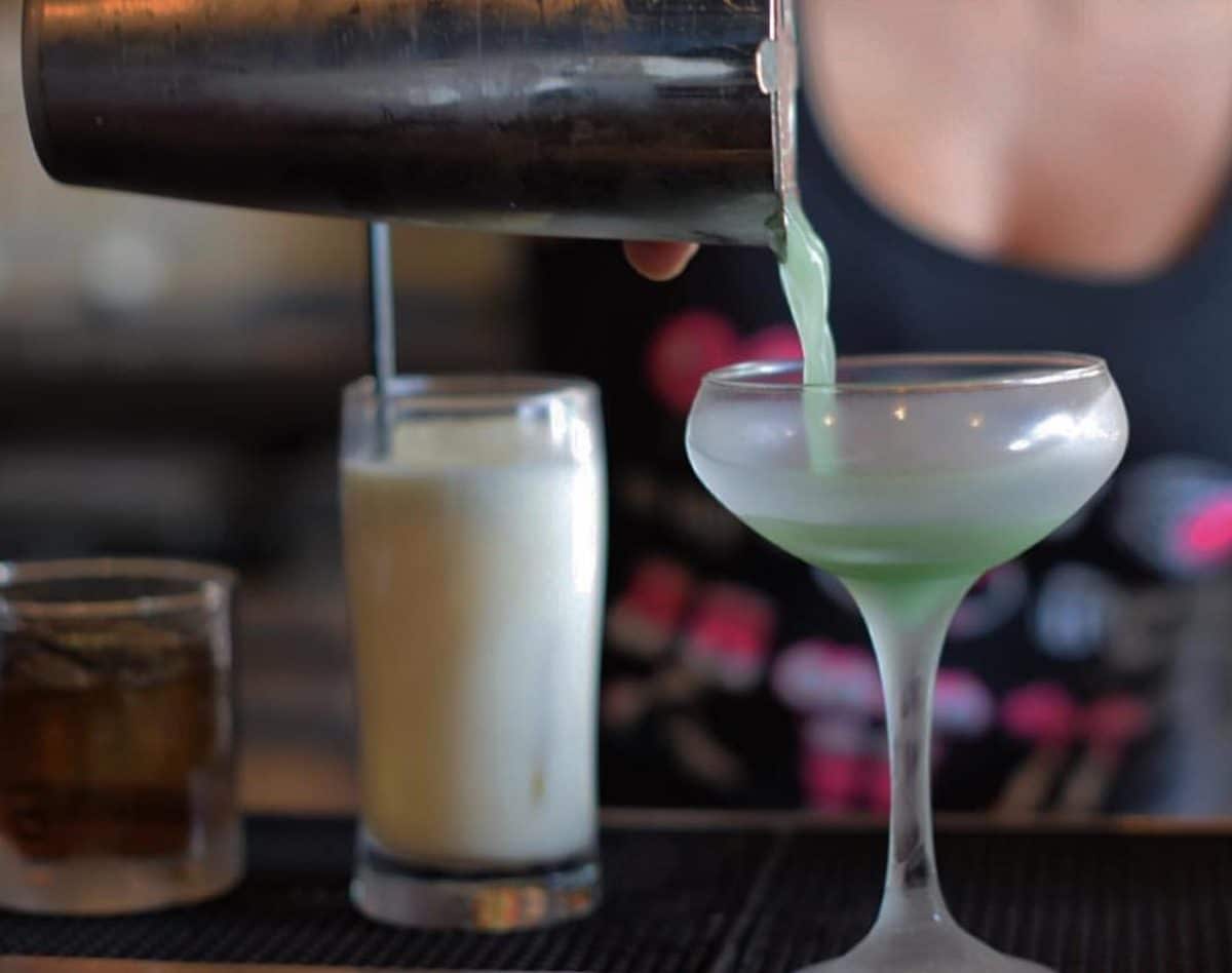 person pouring green drink from mixer into cocktail glass