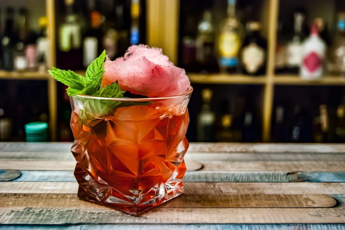 red cocktail with cotton candy and mint sprig on countertop at bar with liquor bottles in blurred background