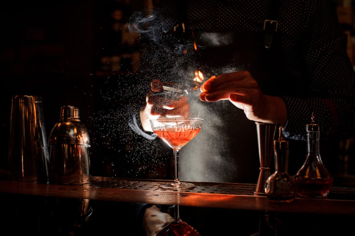 dark bar with bartender lighting a match and burning orange peel for cocktail