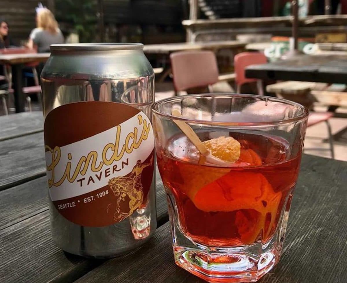 can with sticker reading "linda's tavern" next to a glass both sitting on a table on outdoor patoo