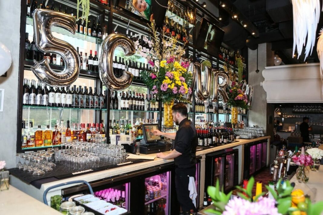 bartender typing into cash register with bottle of liquor on wall above his head and balloons spelling out 5 Church