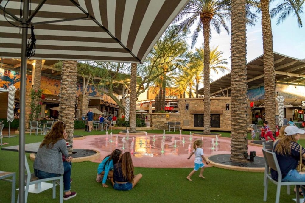 people sitting outside in front of movie theater and shops with splash pad 