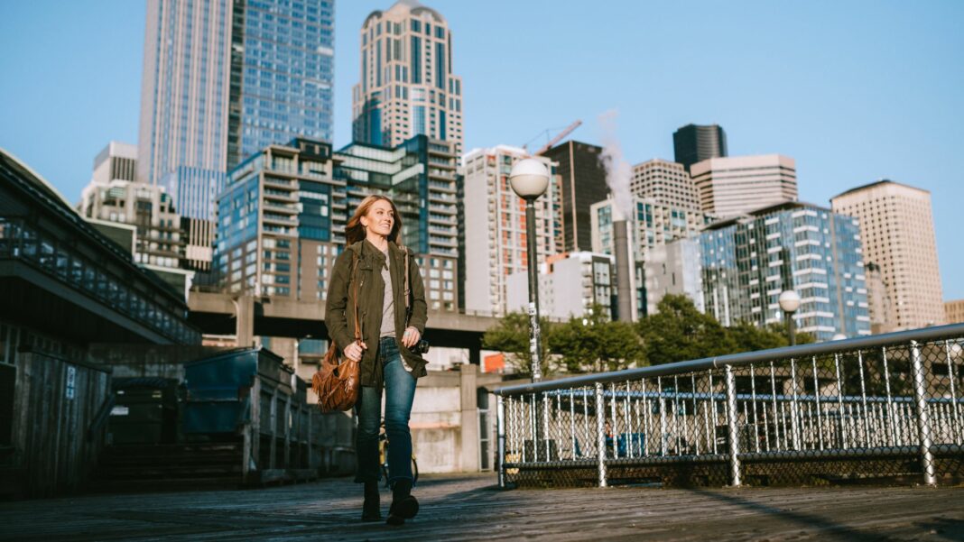 A woman walks on one of the piers of downtown Seattle, the cityscape visible behind her.
